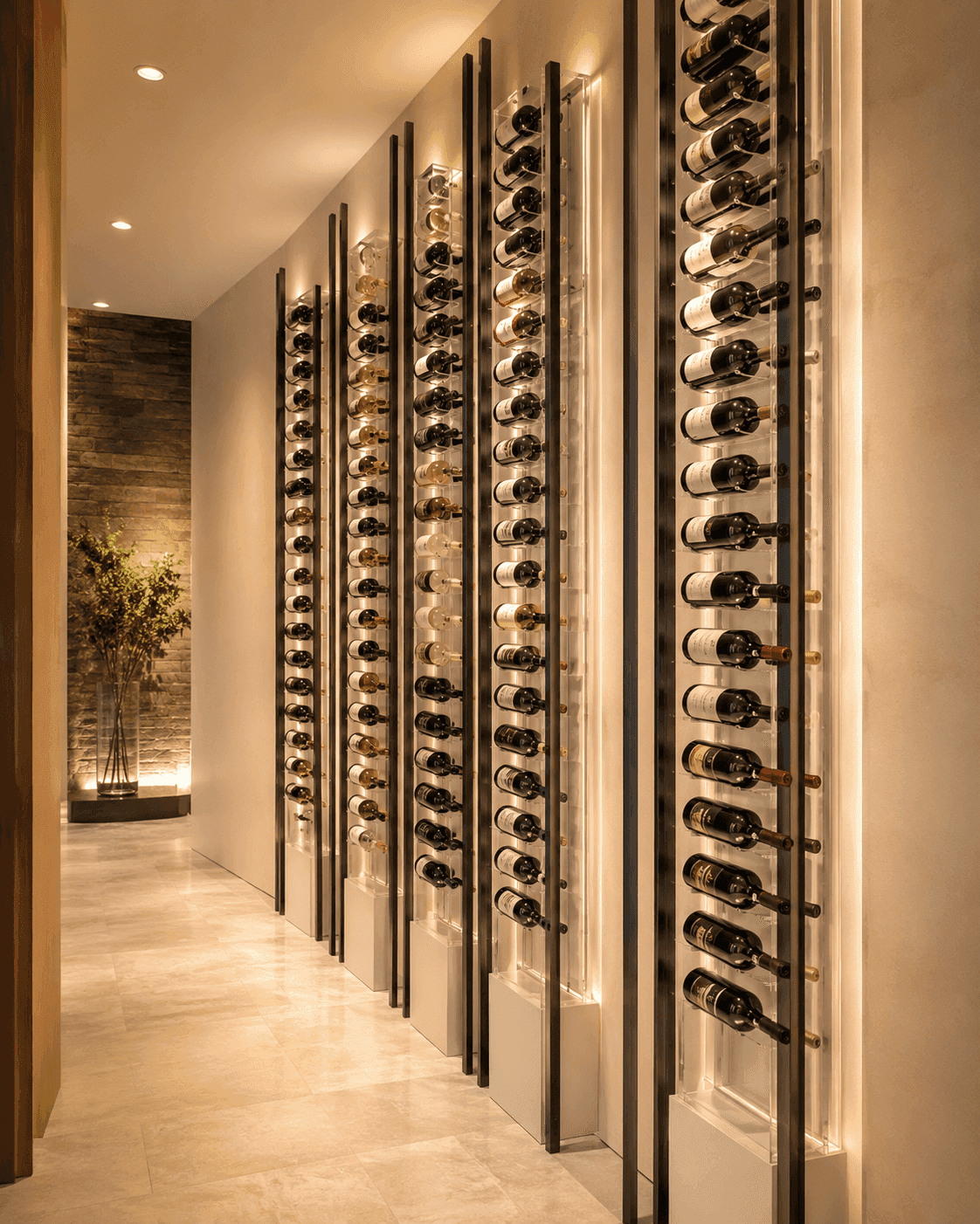 Modern commercial wine cellar with illuminated vertical racks displaying multiple bottles of wine along a hallway with light-colored floors and walls. Tall plant in the background.