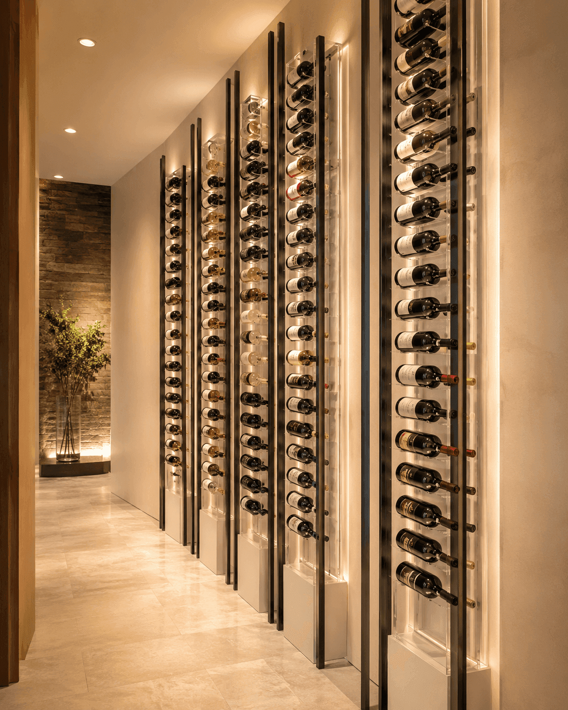 Modern commercial wine cellar with illuminated vertical racks displaying multiple bottles of wine along a hallway with light-colored floors and walls. Tall plant in the background.