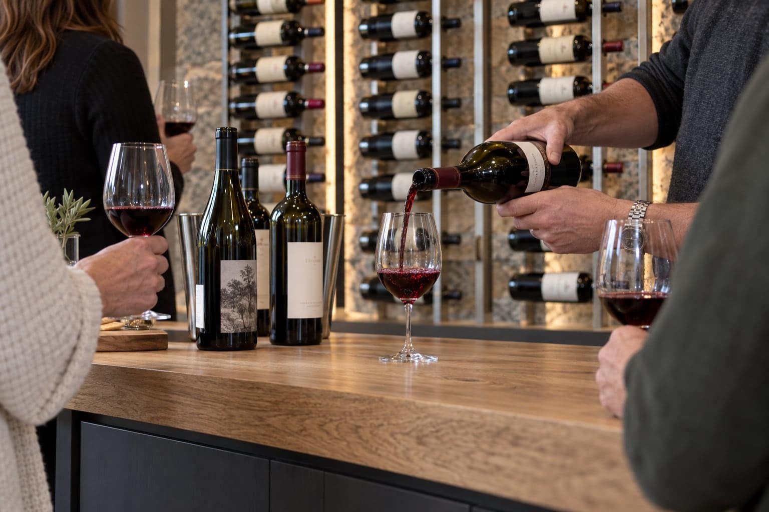 Two people pouring red wine at a counter in front of a lit glass wine wall