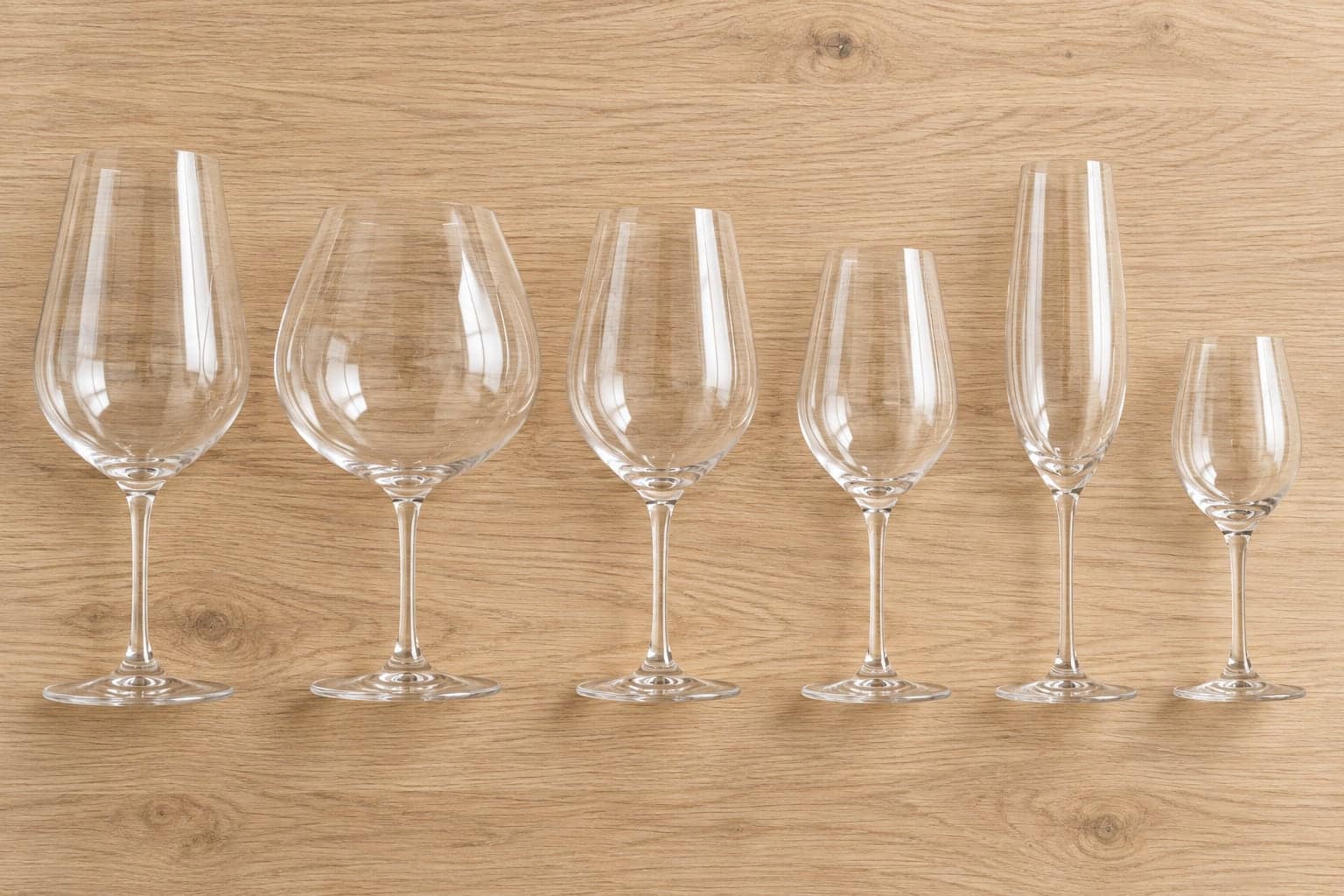 Six empty wine glasses with different bowl shapes arranged on a wooden surface