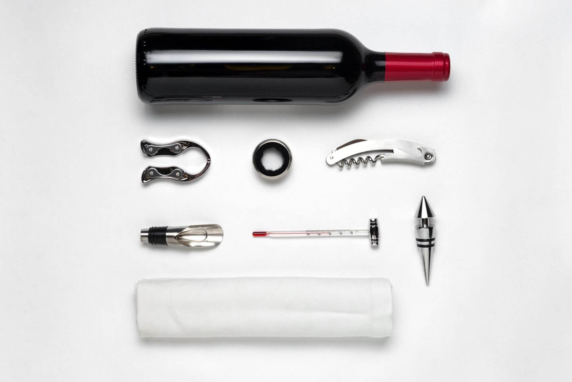 Flat-lay of a wine bottle with foil cutter, corkscrew, stopper, aerator and thermometer on white